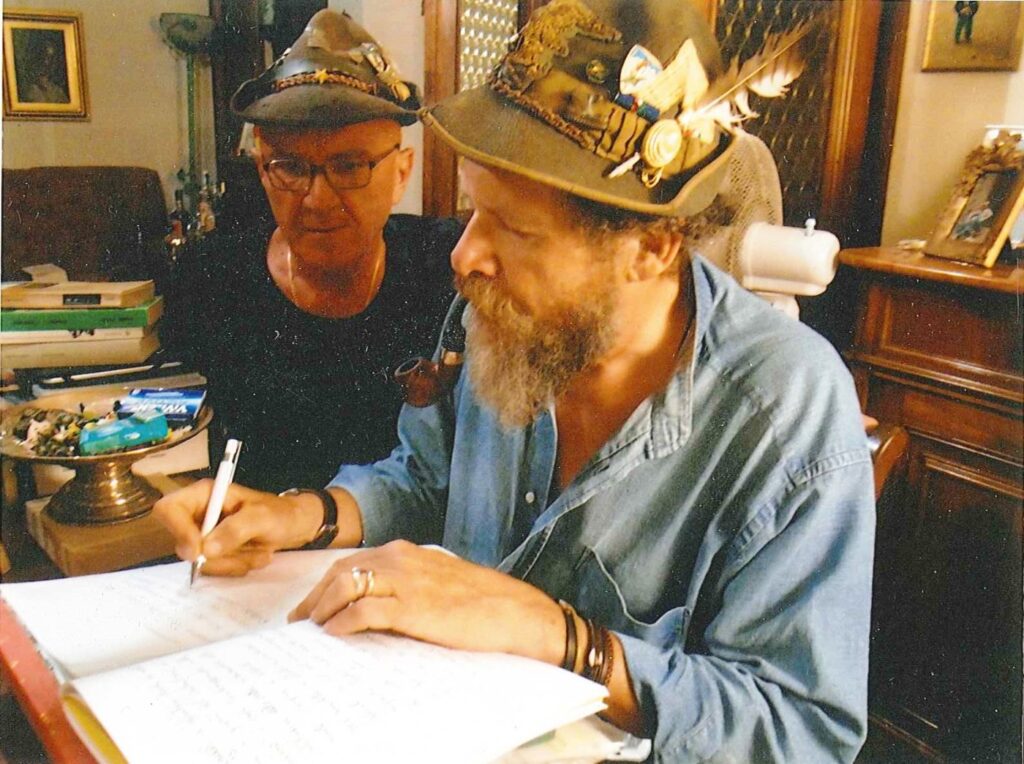 Due uomini seduti a un tavolo: uno, con cappello alpino e barba, scrive su un quaderno