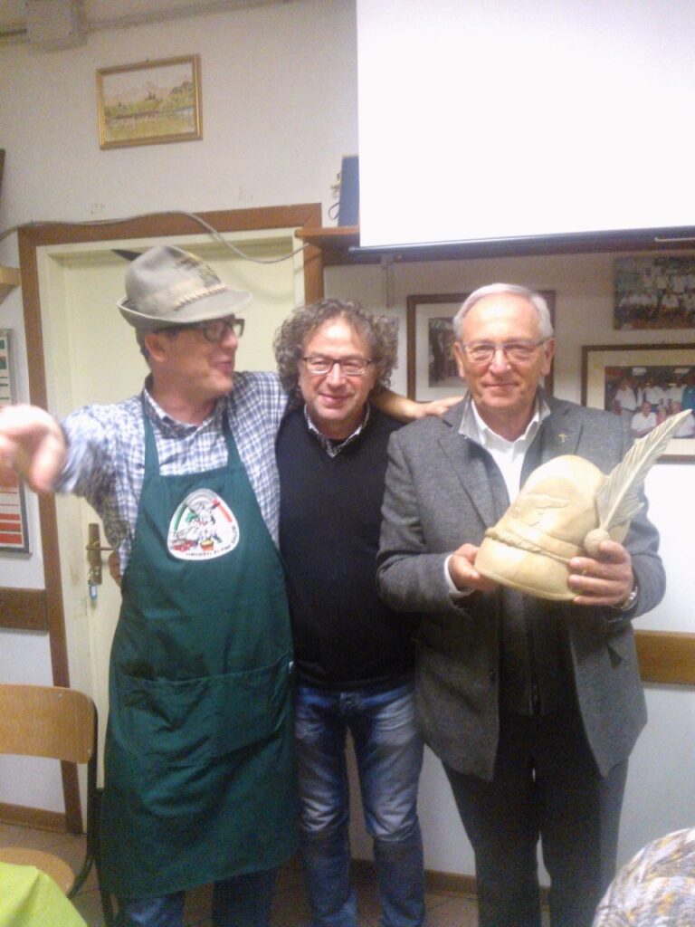 Tre uomini in una sala della sede; uno di loro regge un cappello alpino scolpito nel legno