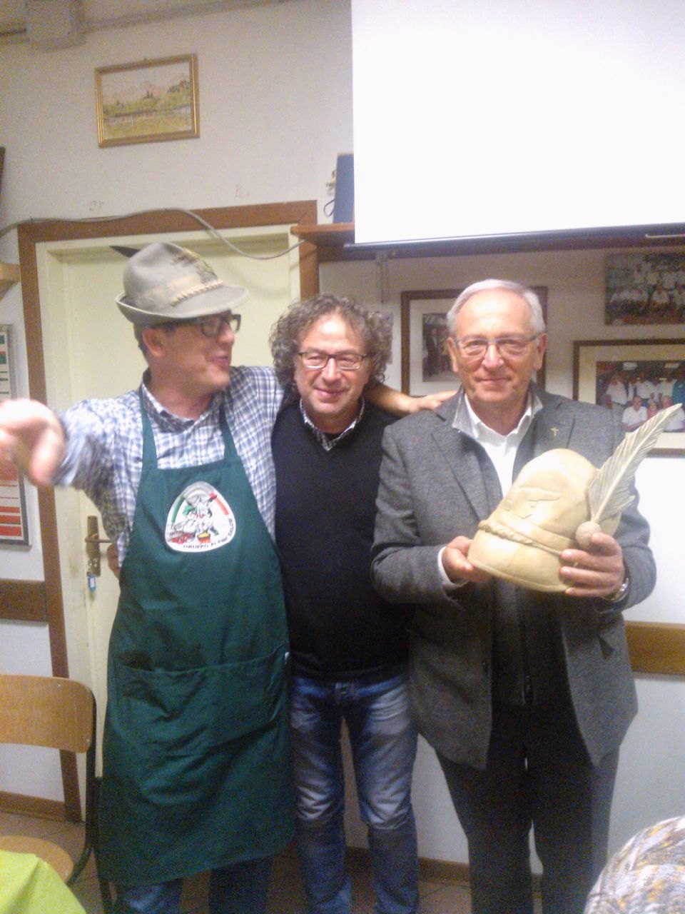 Tre uomini in una sala della sede; uno di loro regge un cappello alpino scolpito nel legno