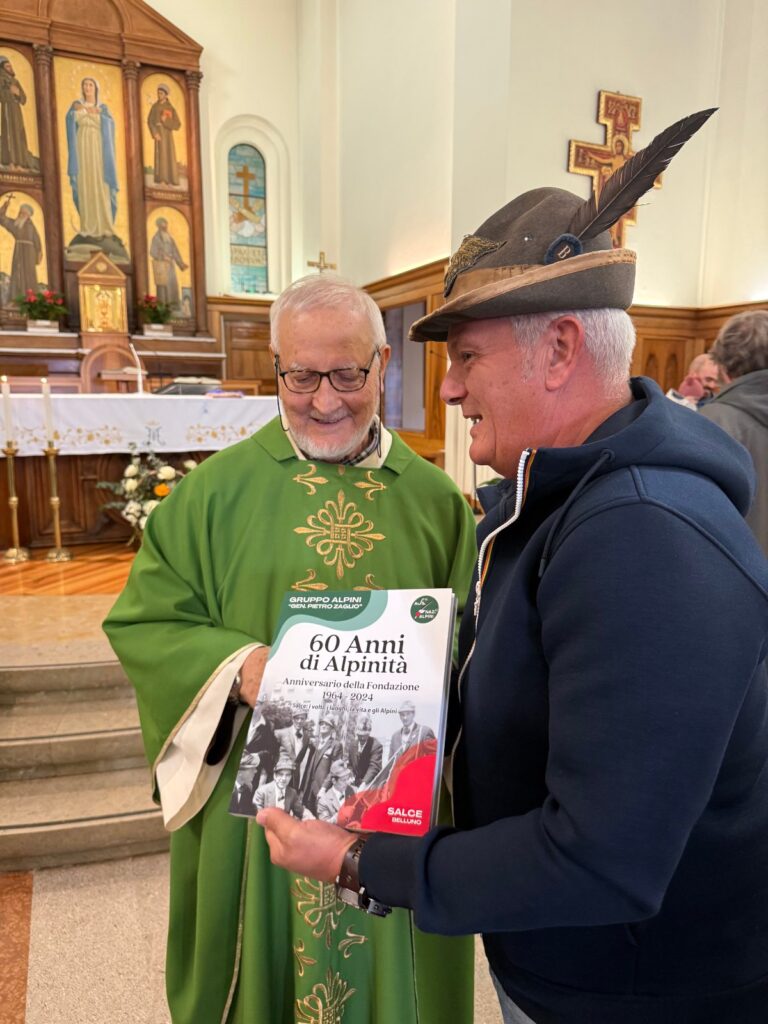 Un sacerdote in paramenti verdi riceve un libro da un alpino con il cappello, davanti all’altare