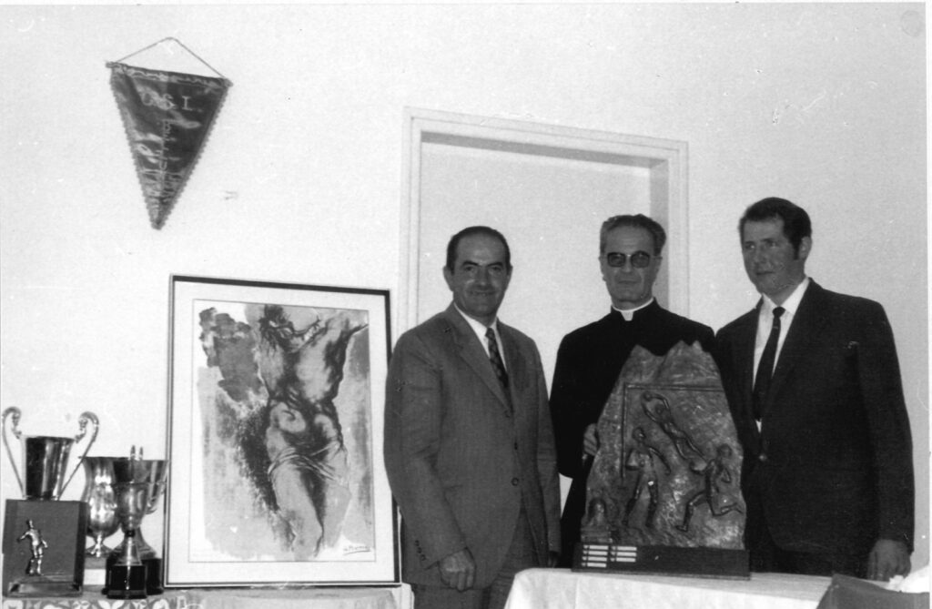 Tre uomini dietro un tavolo con coppe, una scultura e un quadro; alle loro spalle un gagliardetto appeso