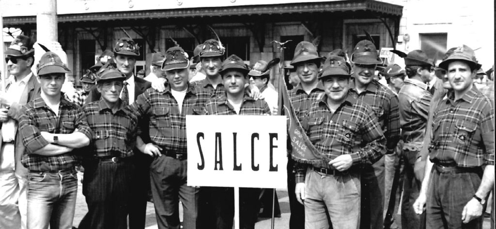Una fila di alpini di Salce in camicia a quadri e cappello alpino, al centro un cartello con la scritta SALCE