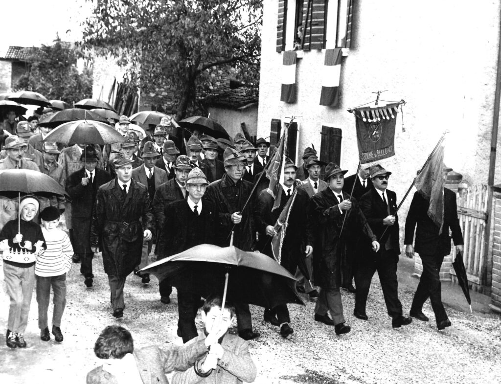 Il corteo degli alpini con gli ombrelli aperti per le strade di Salce il giorno dell’inaugurazione del Gruppo, 1964