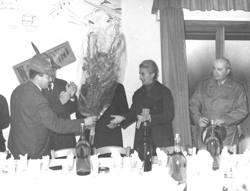 Al pranzo dell'inaugurazione del 1964 un alpino consegna un mazzo di fiori a una signora seduta a tavola