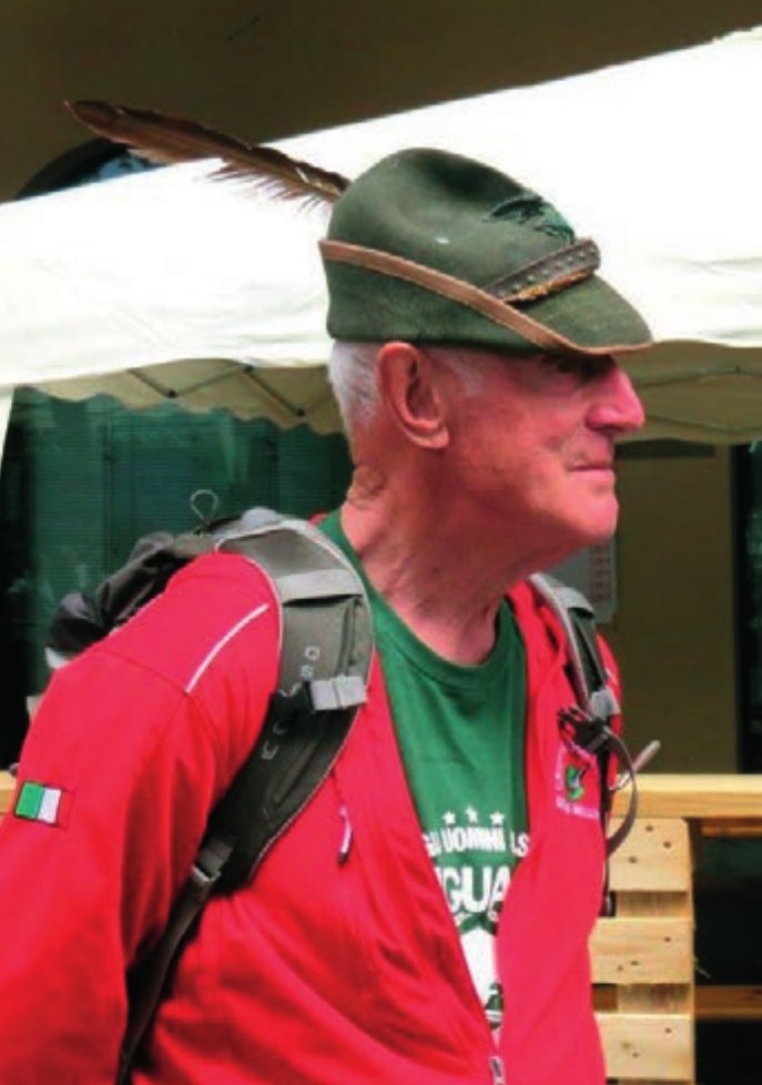 Mario De Luca di profilo, con il cappello alpino con la penna, la giacca rossa del Gruppo e lo zaino in spalla