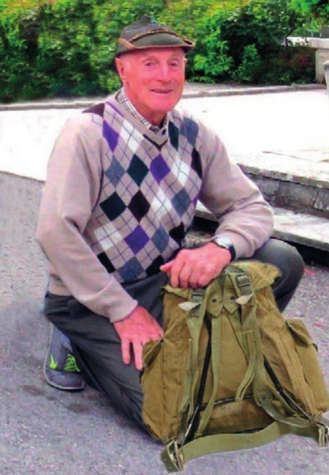 Mario De Luca inginocchiato su una strada di paese, con il cappello e le mani appoggiate sul suo zaino militare verde