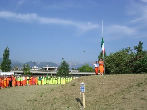 Centinaia di volontari di Protezione Civile in divisa arancione e gialla schierati sul prato durante l’alzabandiera al campo base di Schio