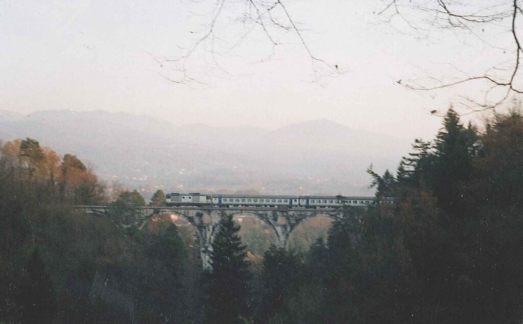 Un treno attraversa il viadotto ad arcate di Val di Siva fra i boschi, con le montagne sullo sfondo