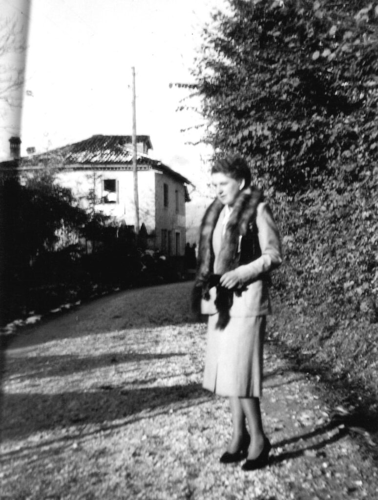 Una giovane donna con cappotto chiaro e collo di pelliccia su una strada di ghiaia; dietro la curva si vede una casa bianca