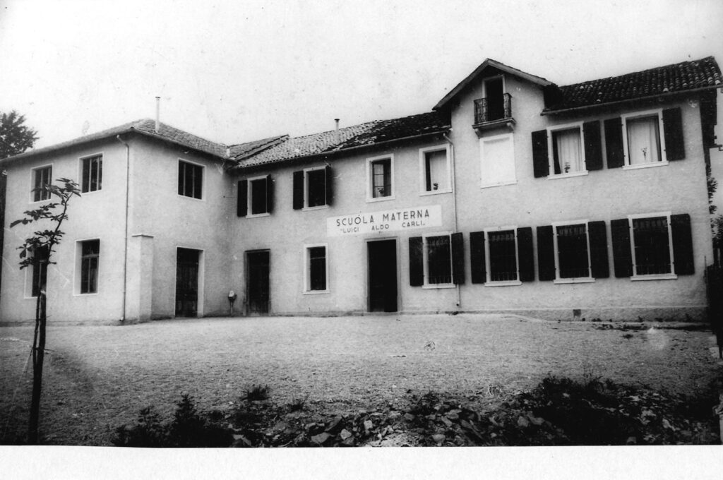 Edificio a due piani con la scritta Scuola Materna sulla facciata e un ampio cortile davanti
