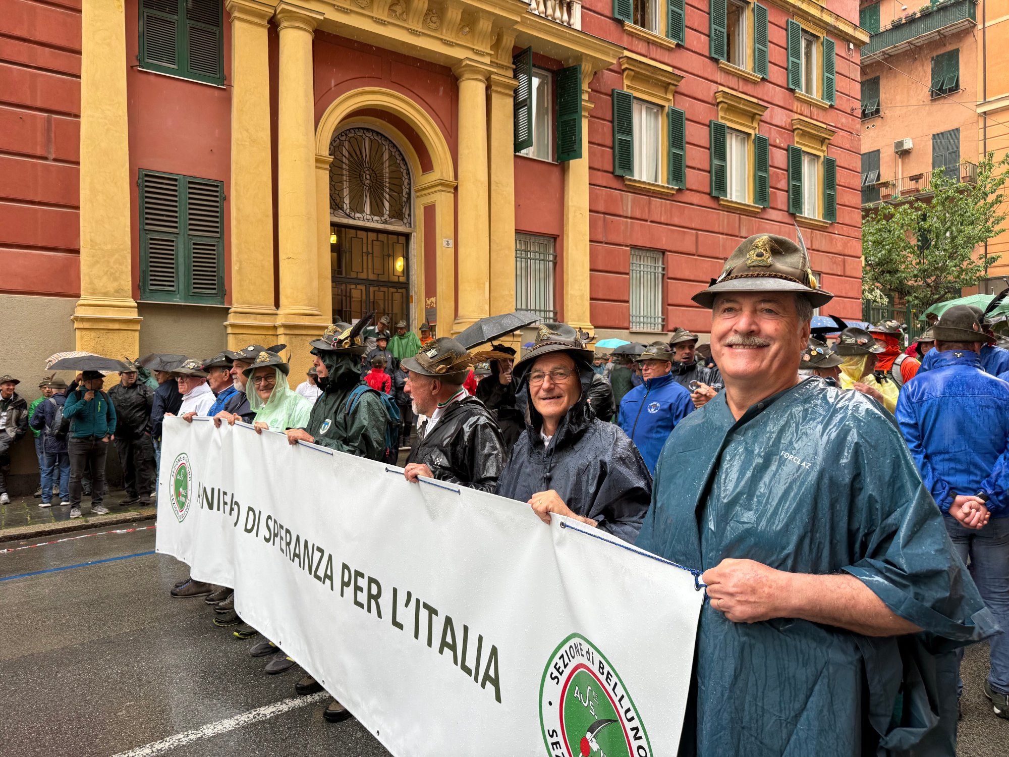 Alpini con il cappello e le mantelle antipioggia reggono lo striscione della Sezione di Belluno in una via di Genova, sotto la pioggia