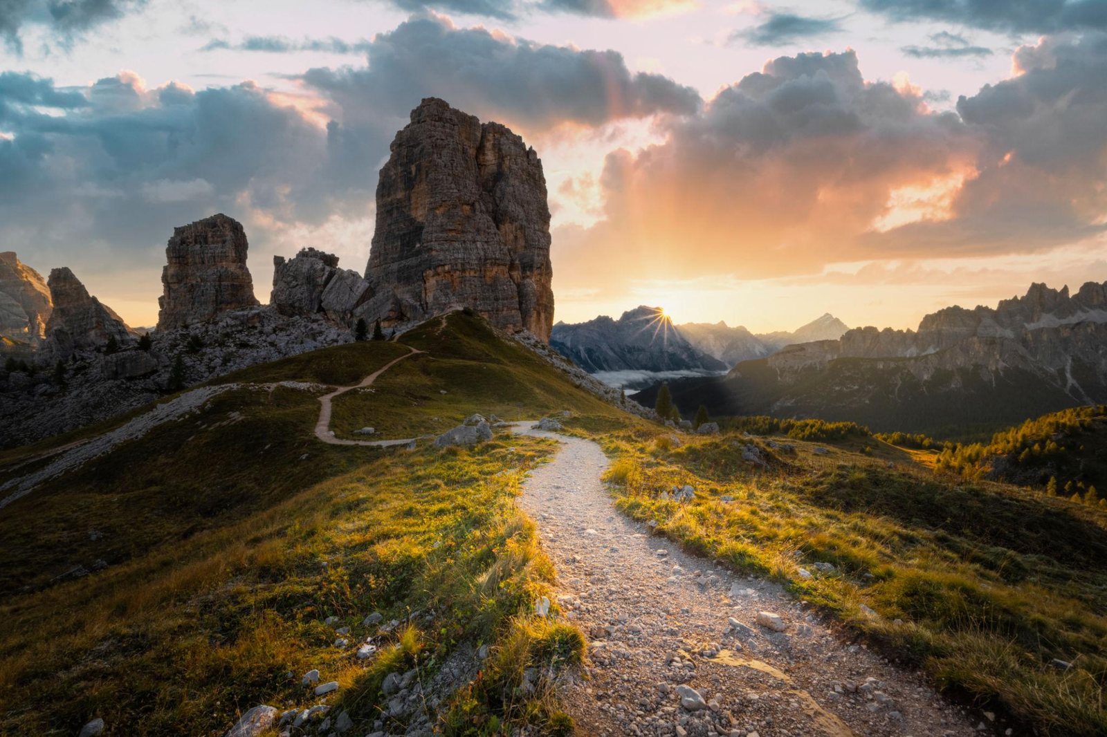 Le Cinque Torri al tramonto con un sentiero di ghiaia tra i prati e il sole che tramonta dietro le montagne