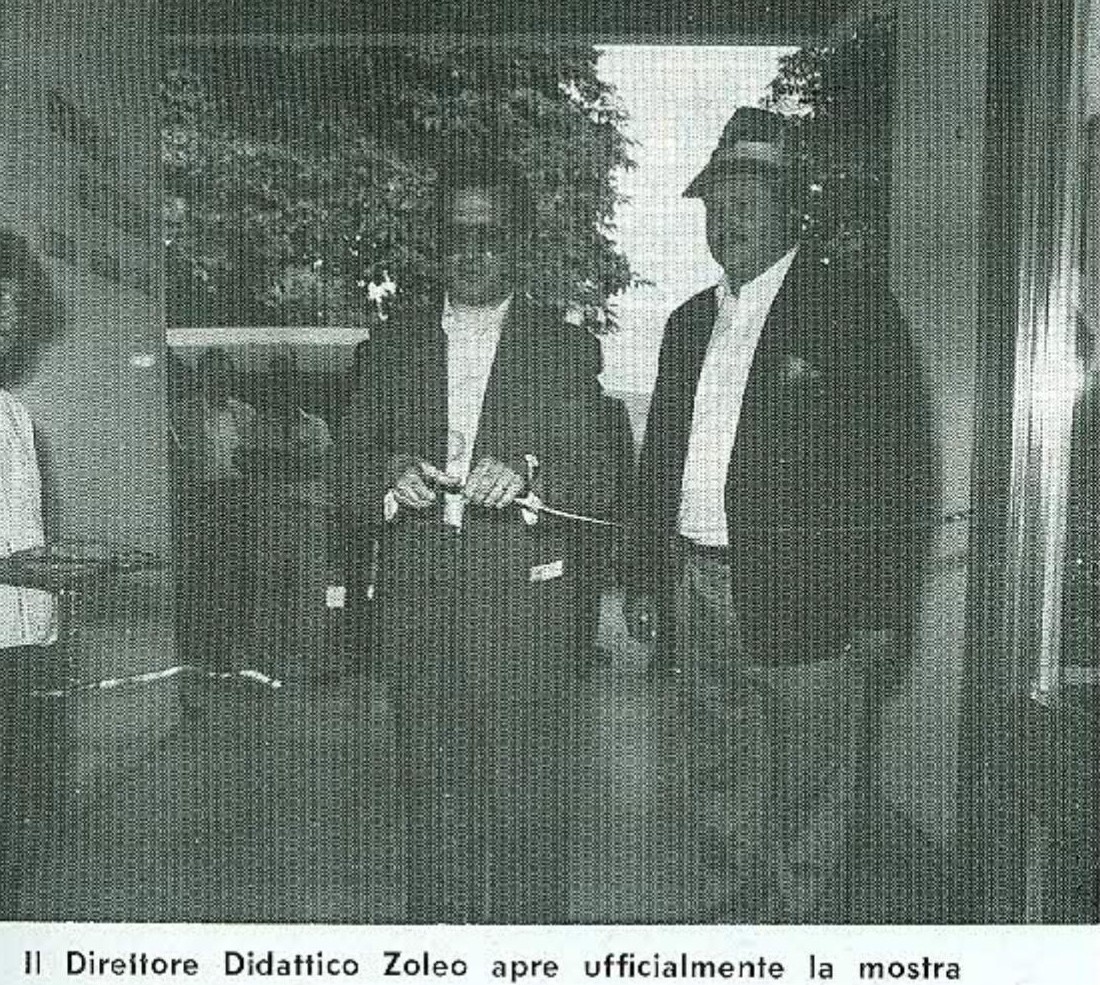 Fotografia in bianco e nero: due uomini davanti a una porta a vetri, uno dei due taglia un nastro