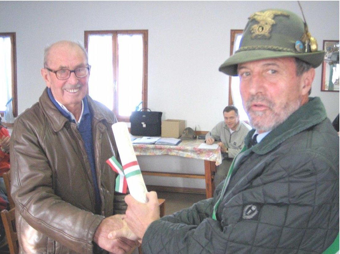 Un anziano con occhiali e giubbotto di pelle stringe la mano a un uomo con il cappello alpino che gli consegna un rotolo con nastro tricolore