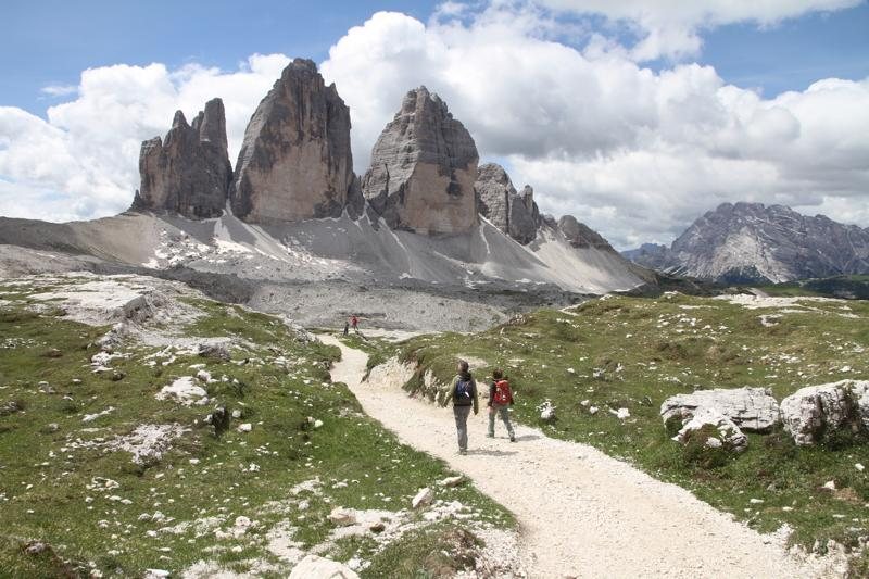 Due escursionisti con lo zaino camminano su un sentiero di ghiaia verso le Tre Cime di Lavaredo sotto un cielo con nuvole bianche